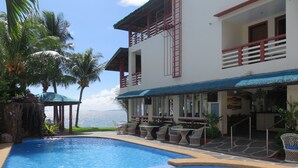 Ein Poolbereich mit Blick auf den Ozean, ein überdachter Sitzbereich und ein Gebäude mit Balkon.