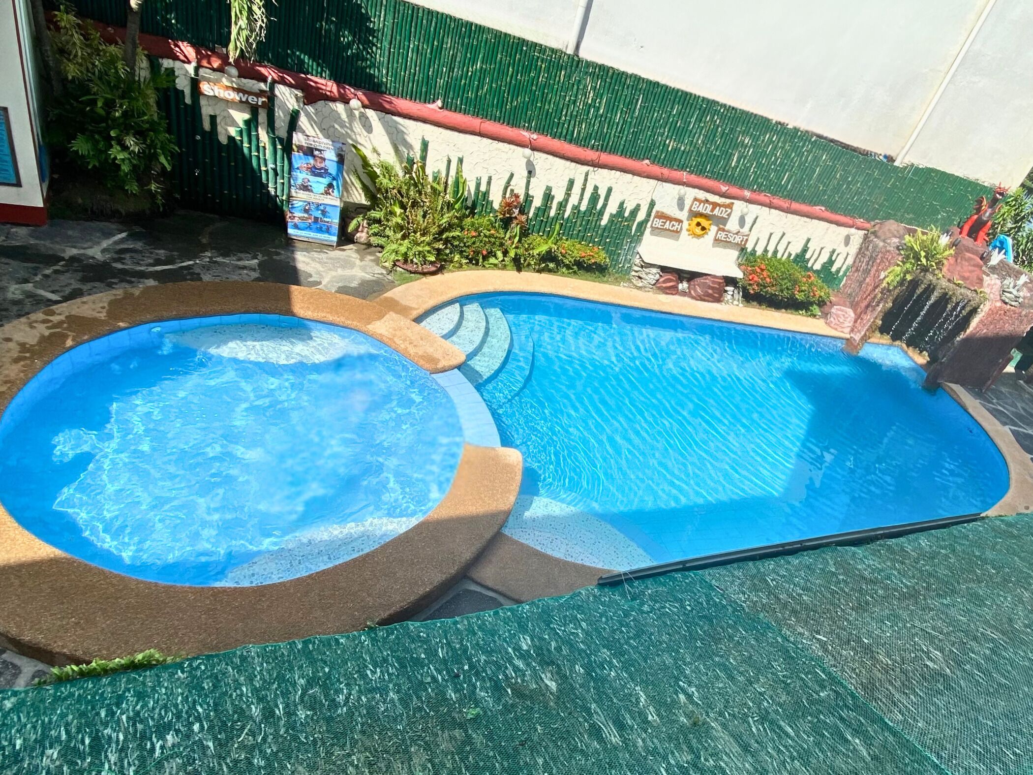 A swimming pool with a hot tub, surrounded by a green fence and a stone wall.