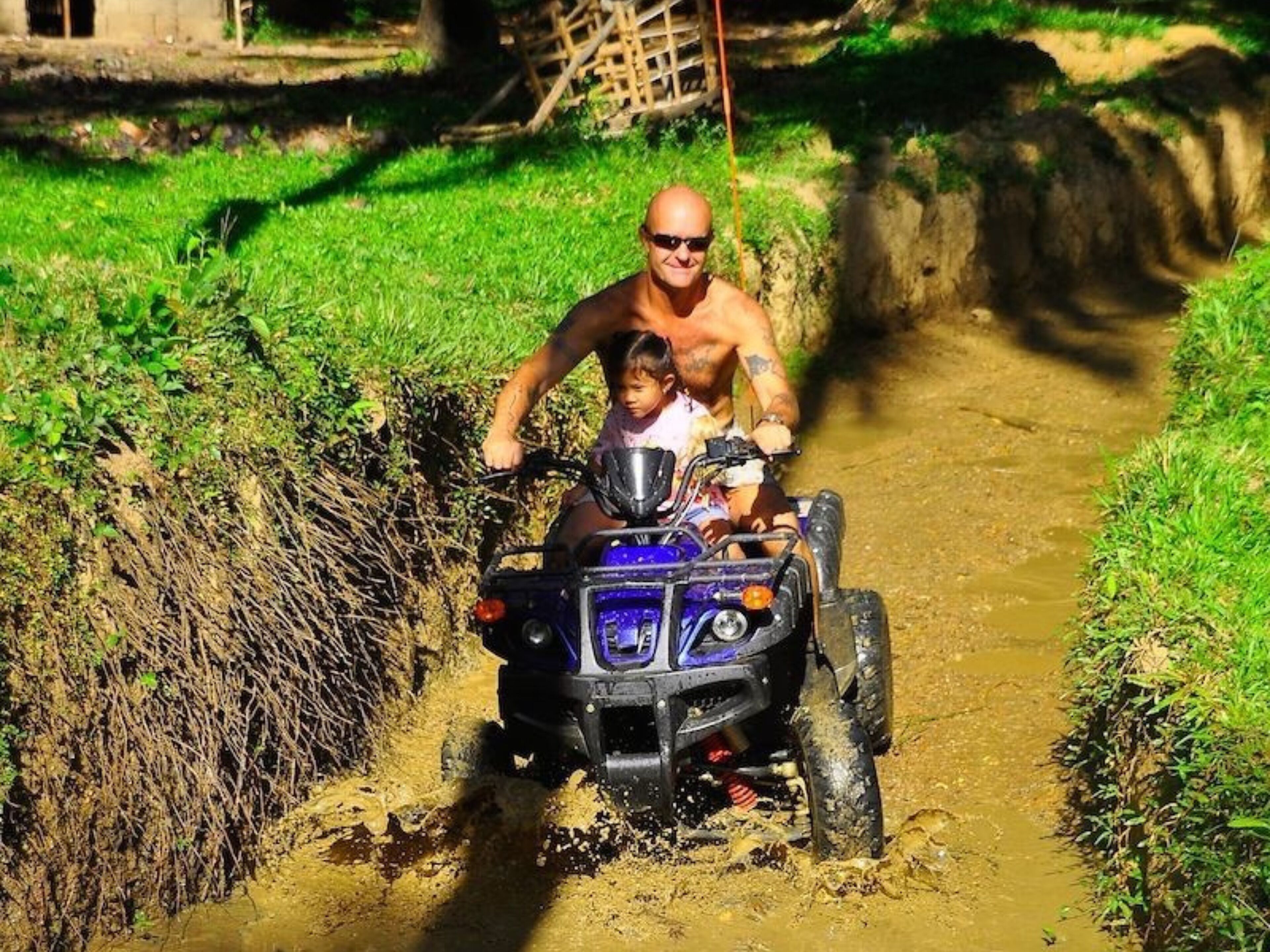 A man and a child on a blue ATV navigating a muddy path.