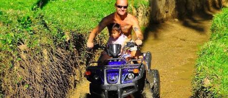 A man and a child on a blue ATV navigating a muddy path.