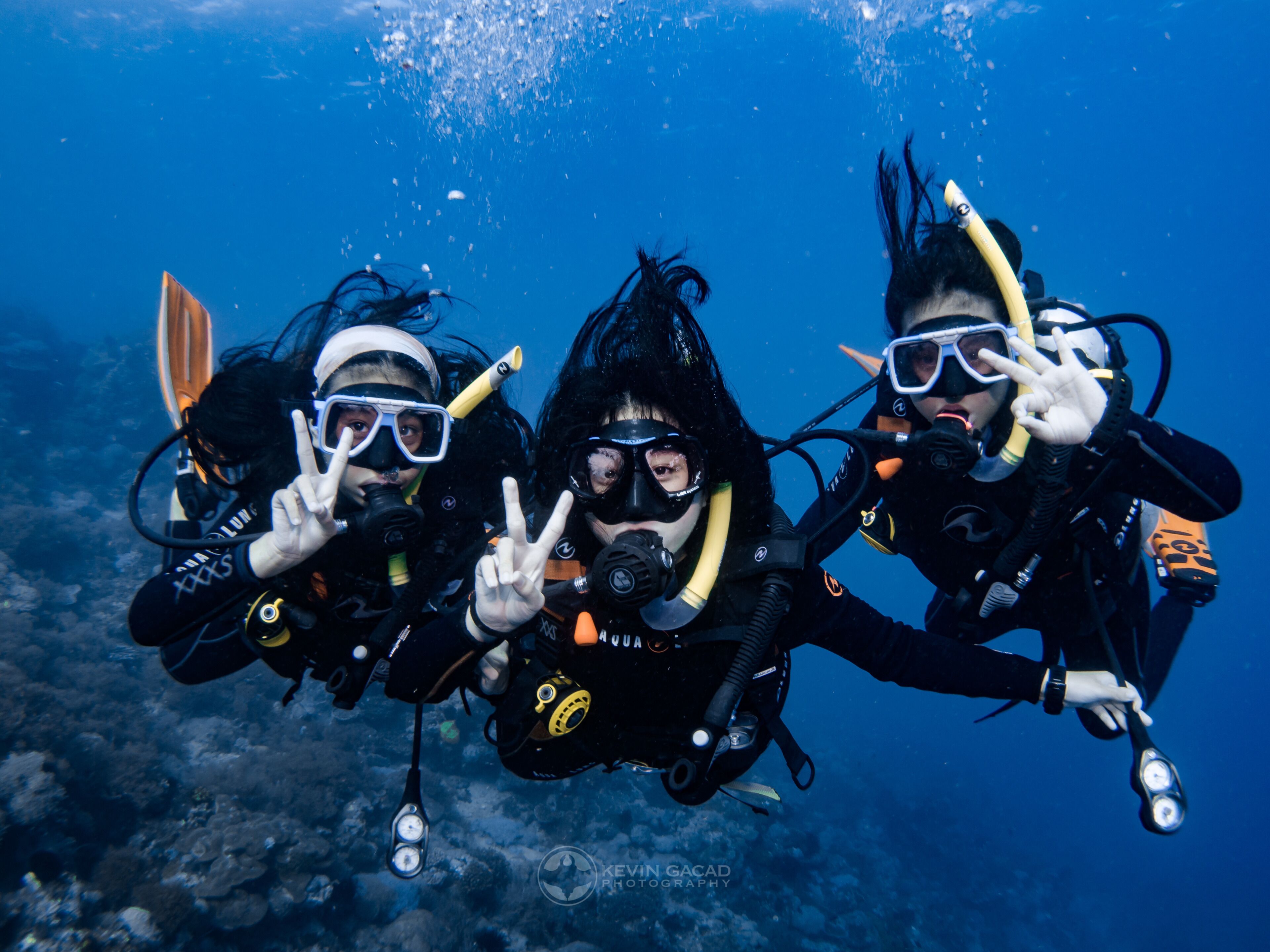 Three scuba divers underwater with scuba gear, making peace signs.