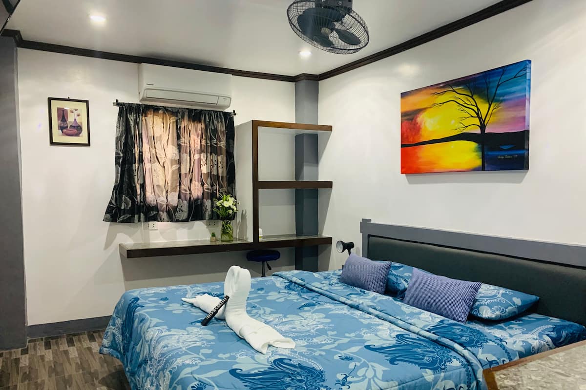 Une chambre à coucher comprenant un lit, une table de chevet, un appareil de climatisation, une fenêtre avec des rideaux, un tableau au mur et un ventilateur.