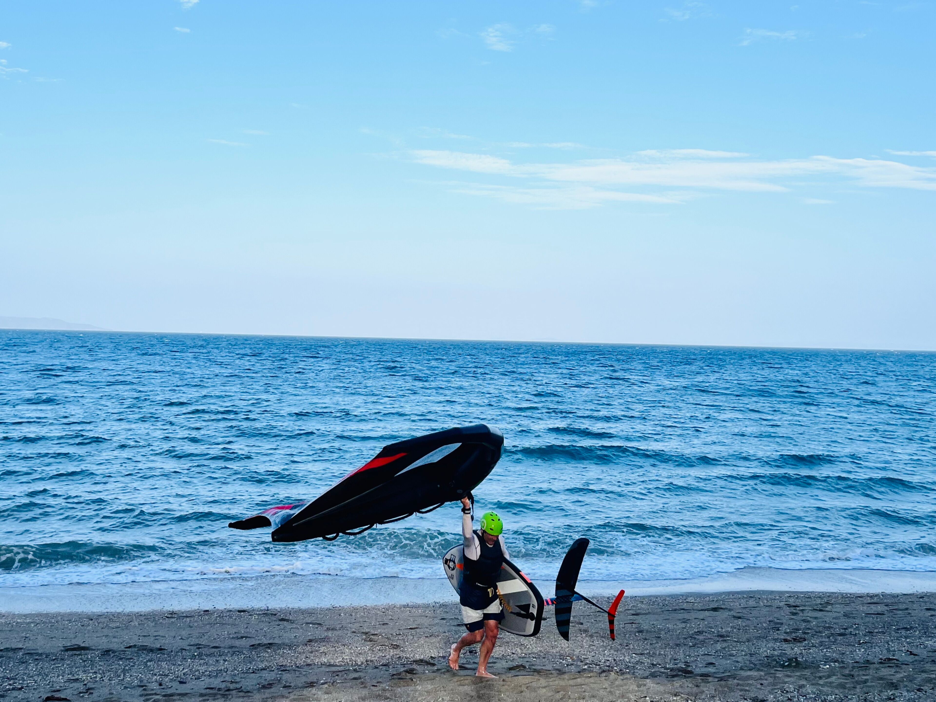 A person carrying a surfboard and a kiteboard on a beach with the ocean in the background.