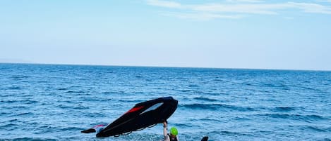 A person carrying a surfboard and a kiteboard on a beach with the ocean in the background.