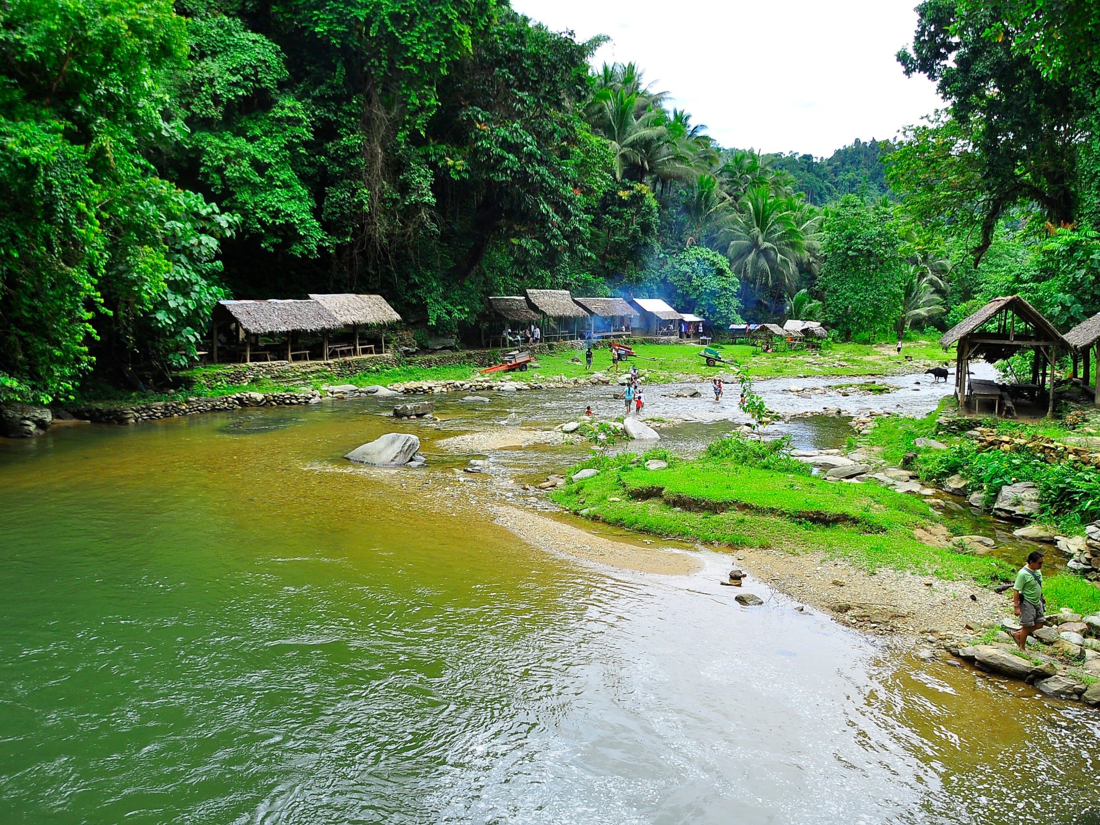A river with rocks and a few people walking along the shore. There are thatched-roof huts and lush greenery in the background.