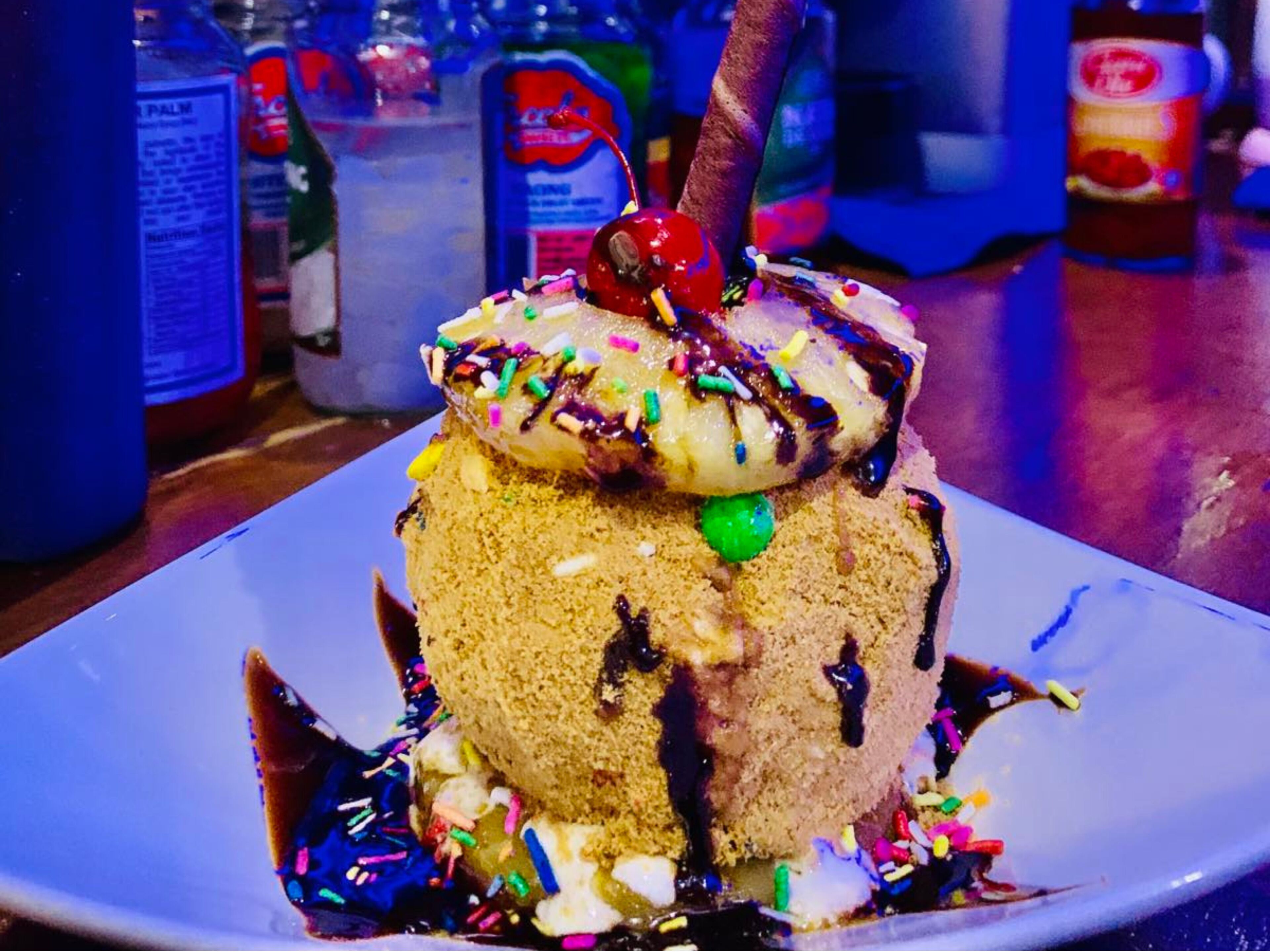 A dessert on a plate with a cherry on top, surrounded by colorful sprinkles and chocolate sauce.