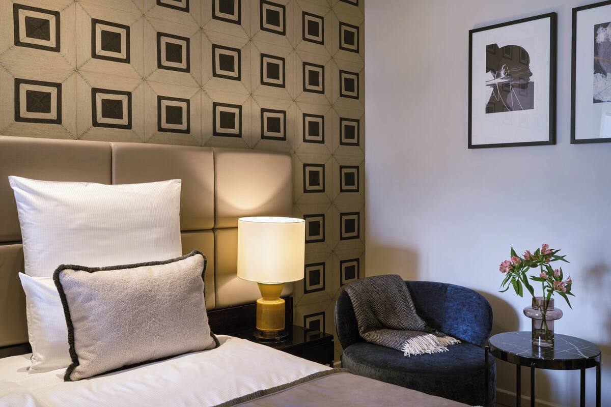 Ein Hotelzimmer mit einem Bett, einem Nachttisch mit Lampe, einem Sessel und einem kleinen Tisch mit Blumenvase.