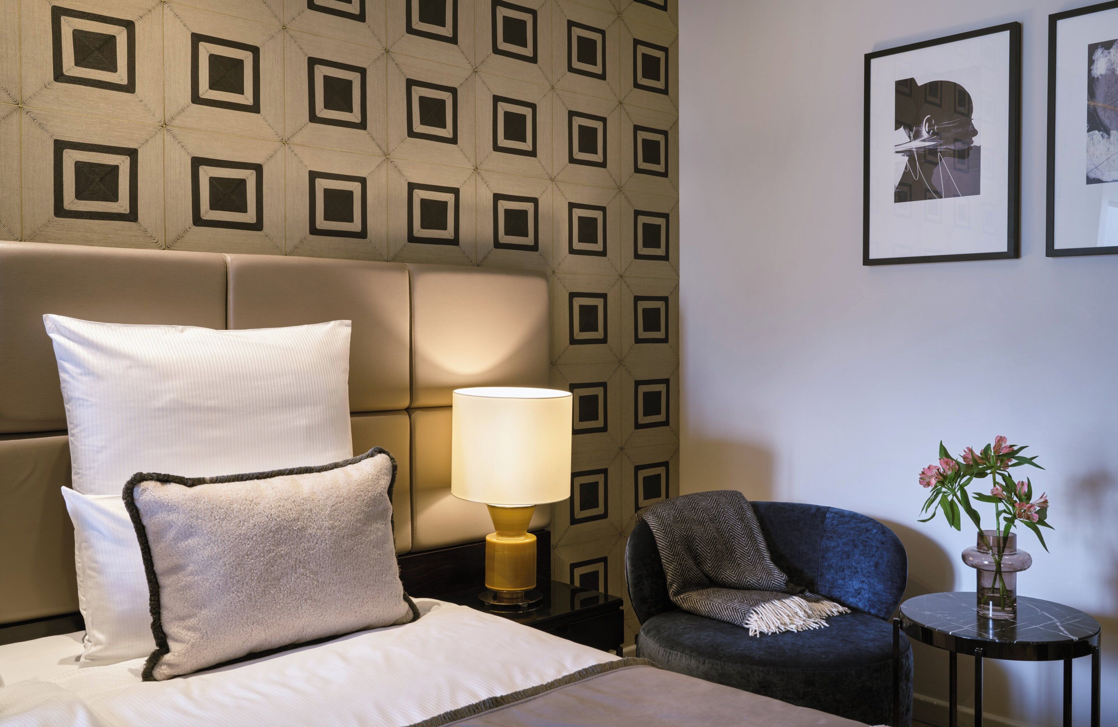 Habitación de hotel con cama, mesita de noche con lámpara, una silla y una pequeña mesa con un jarrón de flores.