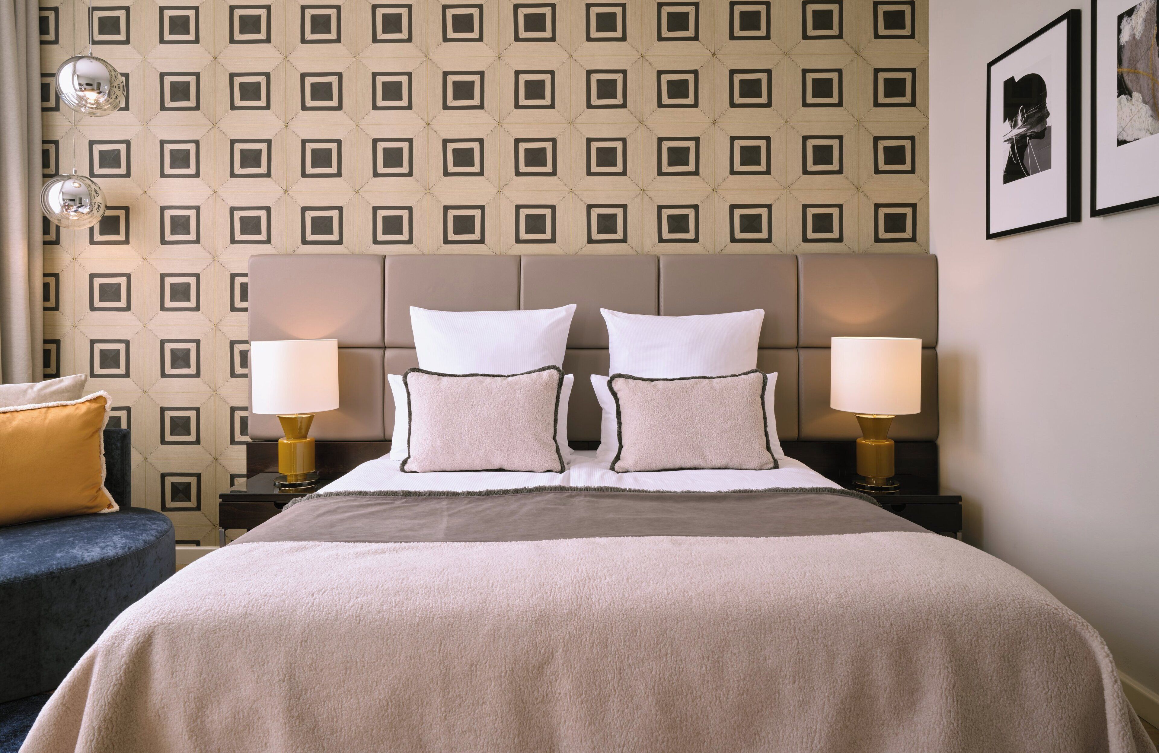 Une chambre d’hôtel avec un papier peint à motifs, un grand lit avec deux oreillers blancs, deux lampes de chevet et un fauteuil bleu.