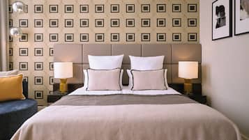 Une chambre d’hôtel avec un papier peint à motifs, un grand lit avec deux oreillers blancs, deux lampes de chevet et un fauteuil bleu.