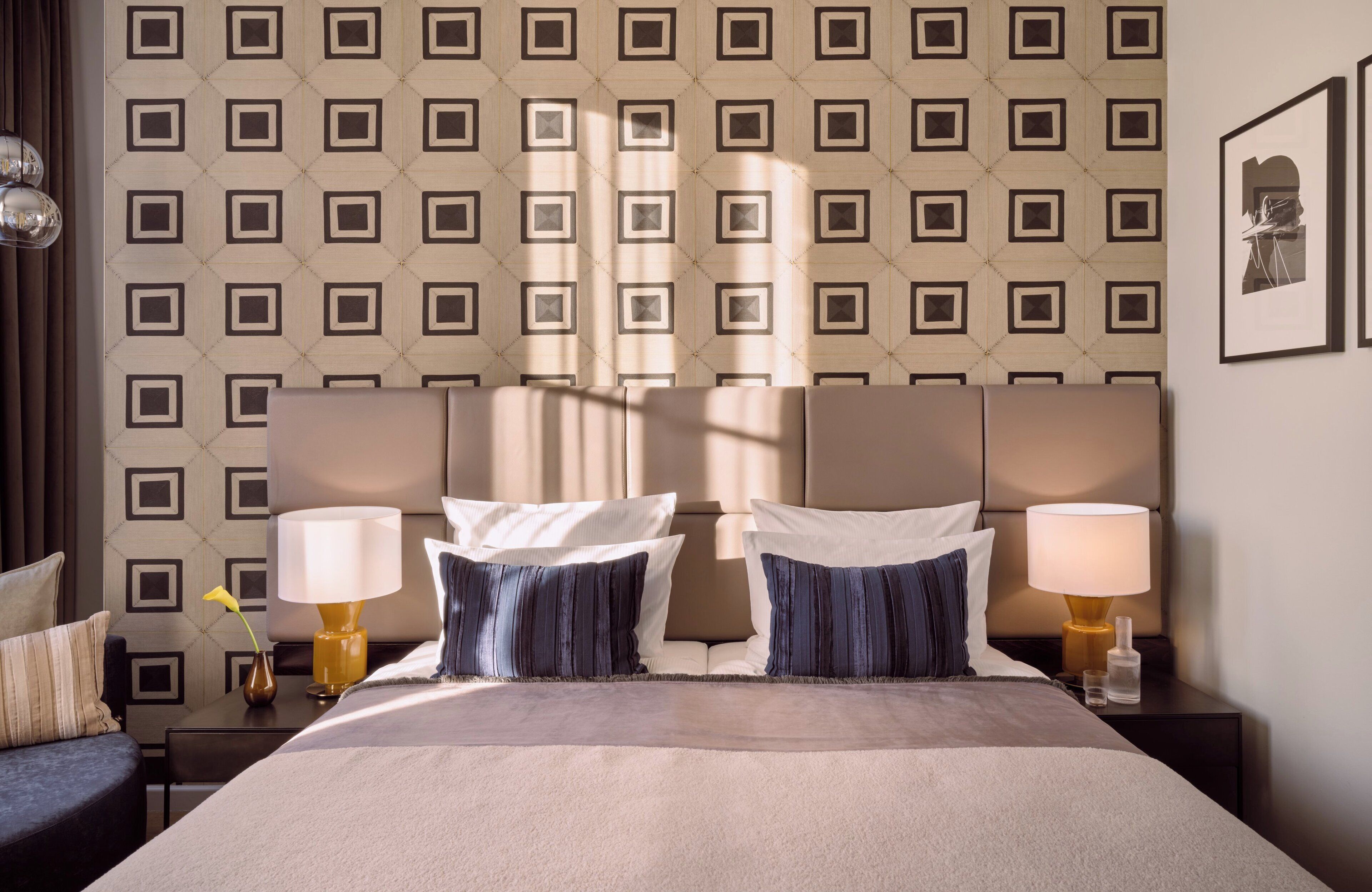A hotel room with a large bed, two bedside tables with lamps, a patterned wallpaper, and a framed picture on the wall.