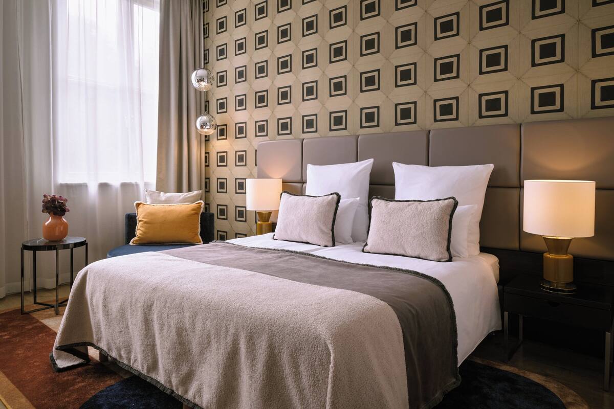 Ein ordentlich eingerichtetes Hotelzimmer mit einem großen Bett, zwei Nachttischen, einer Lampe und einer Blumenvase, sowie einer Wand mit Muster.