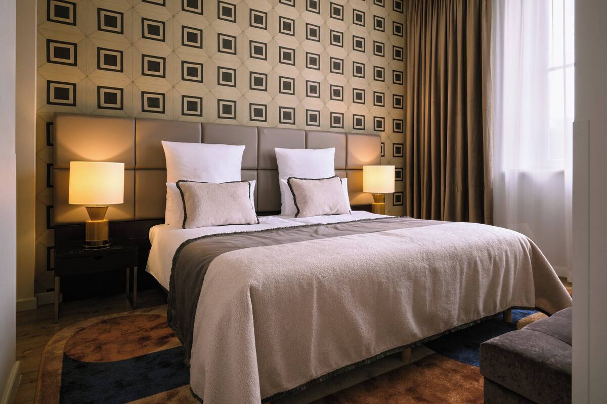 Ein Hotelzimmer mit einem großen Bett, zwei Nachttischlampen, einer Wand mit Muster und einem Vorhangfenster.