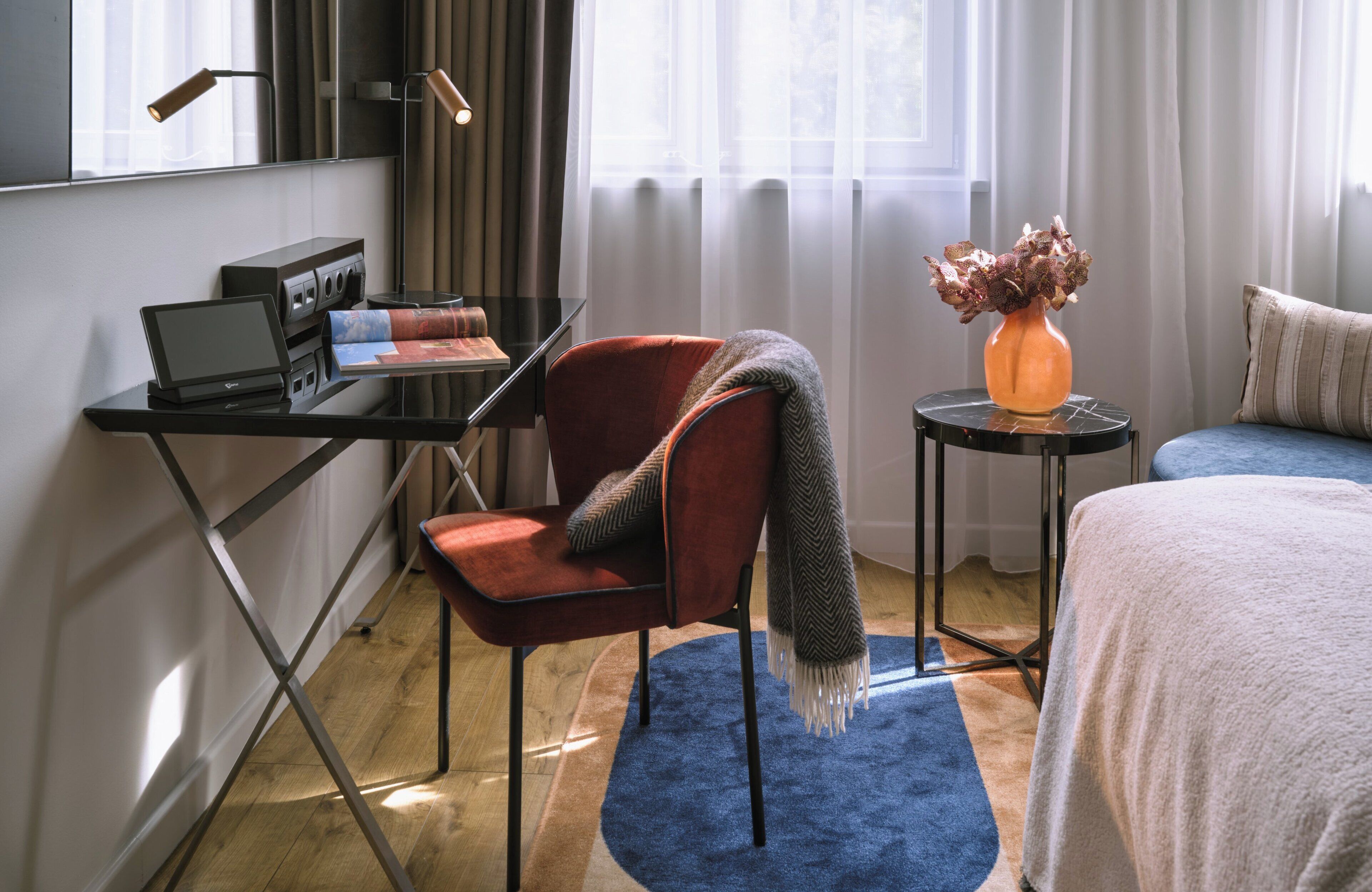 Une chambre d’hôtel moderne avec un bureau, un ordinateur portable et une chaise rouge. On y trouve une petite table sur laquelle est posé un vase rempli de fleurs séchées, ainsi qu’un tapis bleu.