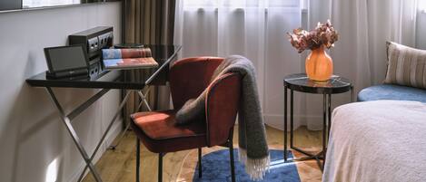 A modern hotel room with a desk, laptop, and a red chair. There is a small table with a vase of dried flowers and a blue rug.