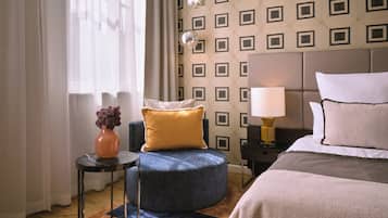 A hotel room with a bed, a round blue ottoman, a small table with a vase, and a patterned wall.