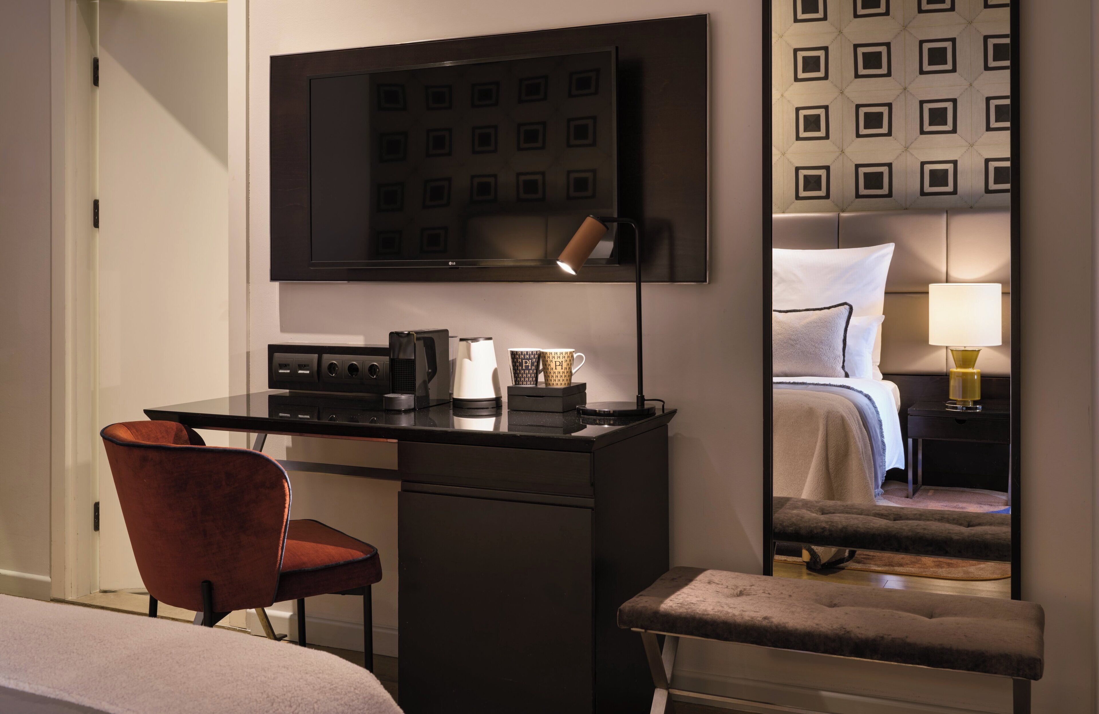 A hotel room with a desk, a chair, a bed with a headboard, a bedside table with a lamp, and a television mounted on the wall.