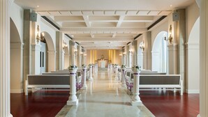 A long, narrow hallway with rows of white benches and columns, leading to a central area with a chandelier.