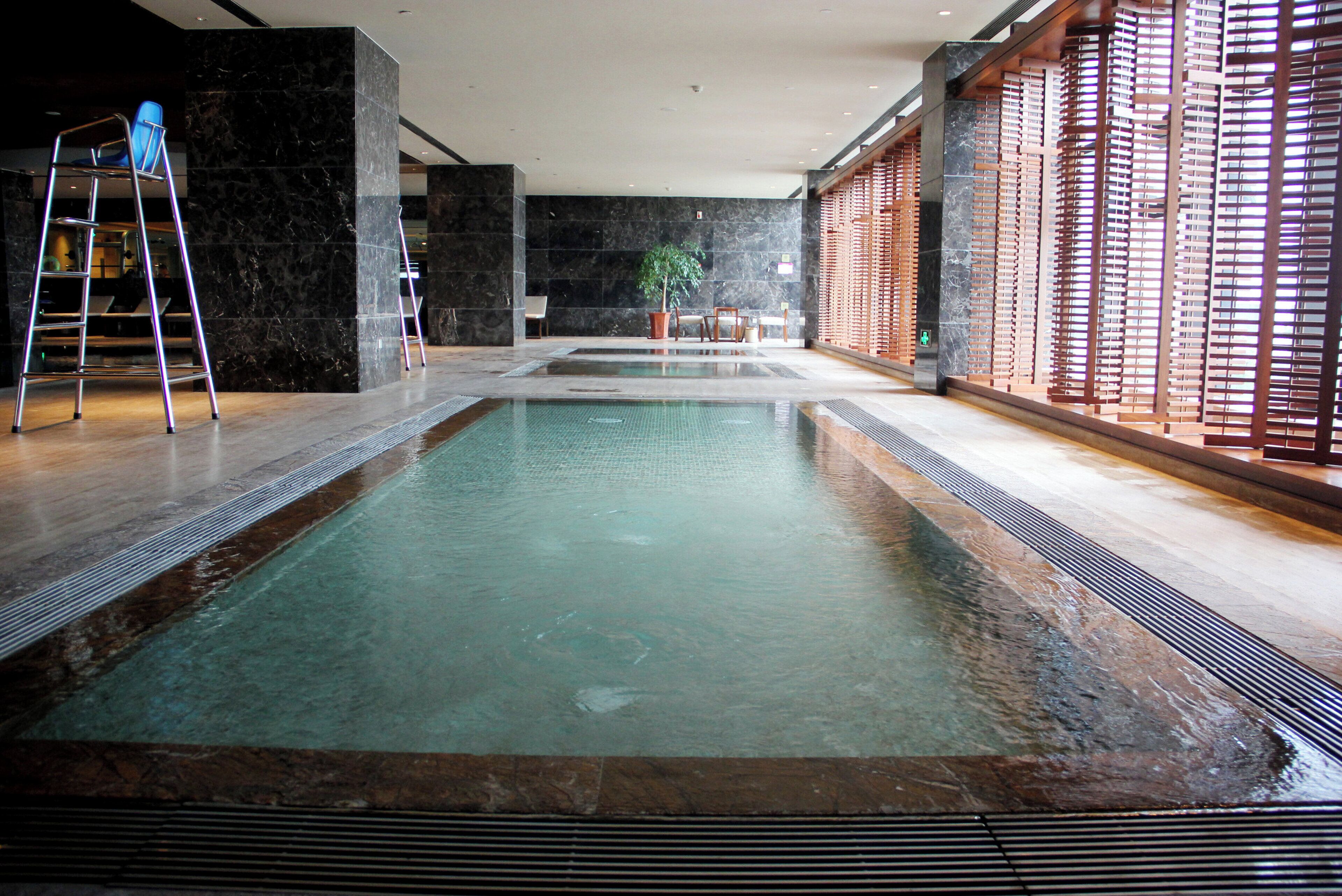 An indoor swimming pool with a clear water surface, surrounded by marble walls and wooden flooring.
