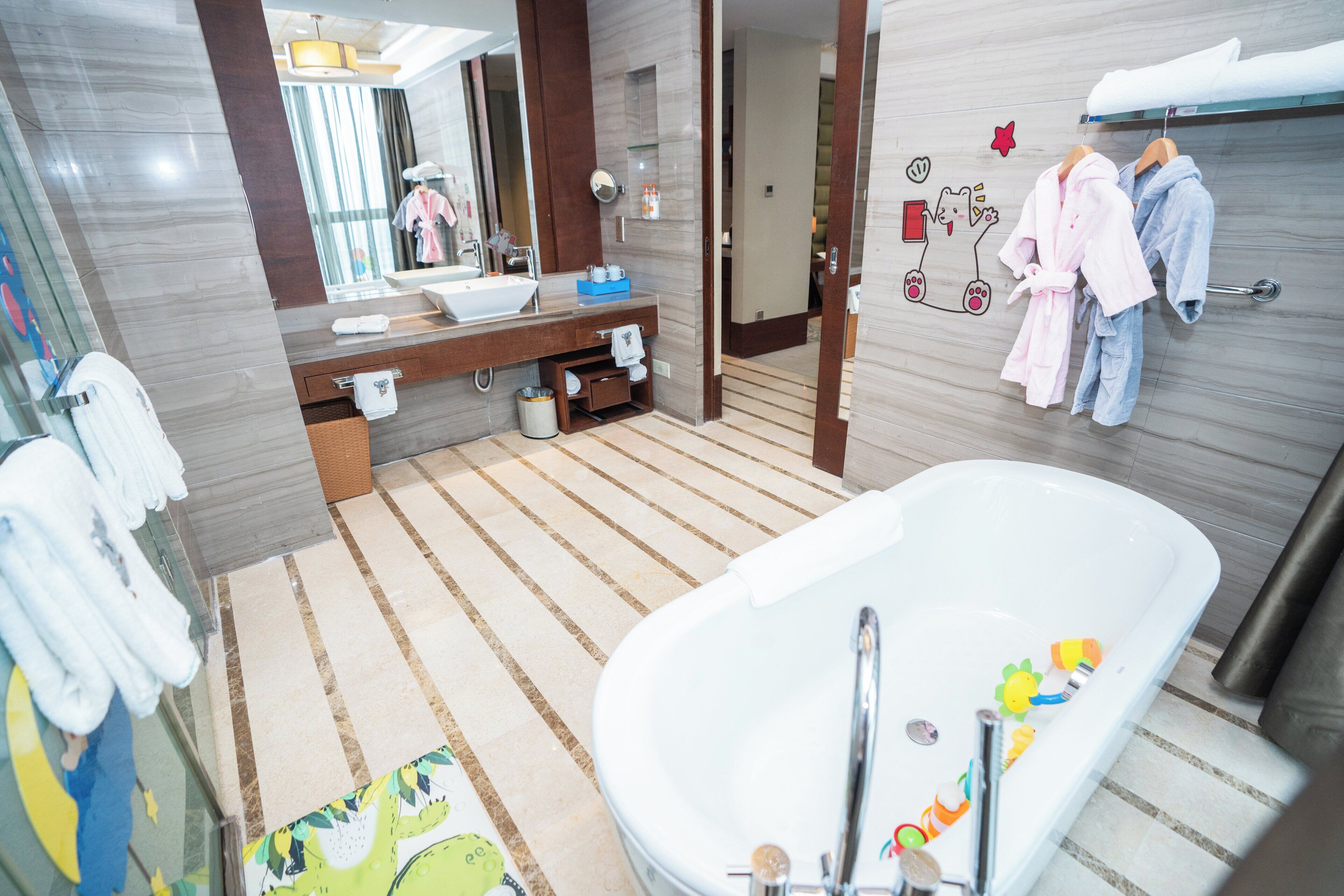 A modern bathroom with a large bathtub, a vanity with a sink, and a mirror. There are towels and robes hanging on a rack, and a small trash bin is visible.