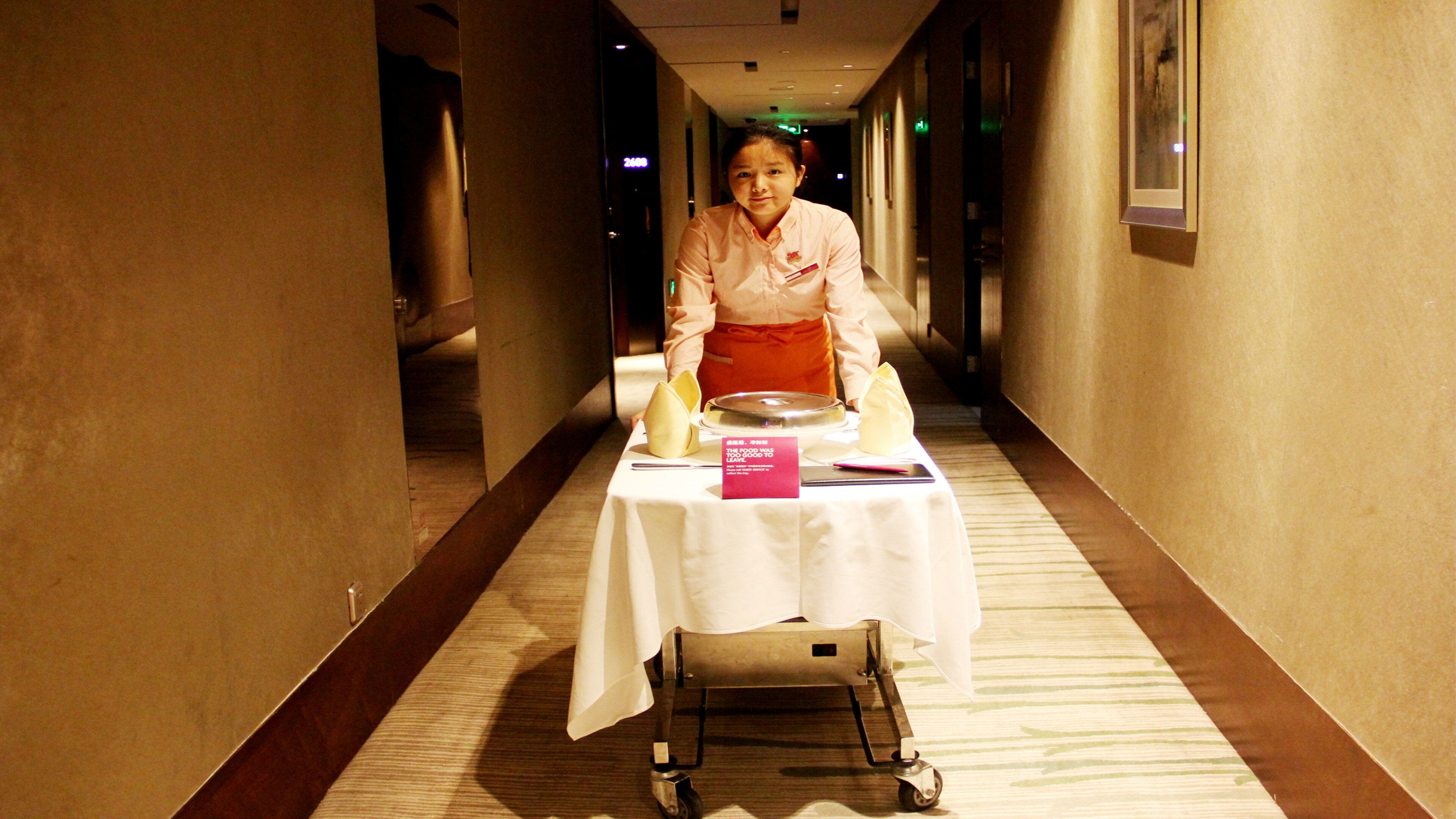 A hotel corridor with a table set for a meal, a sign indicating the food was left, and a person in an apron.