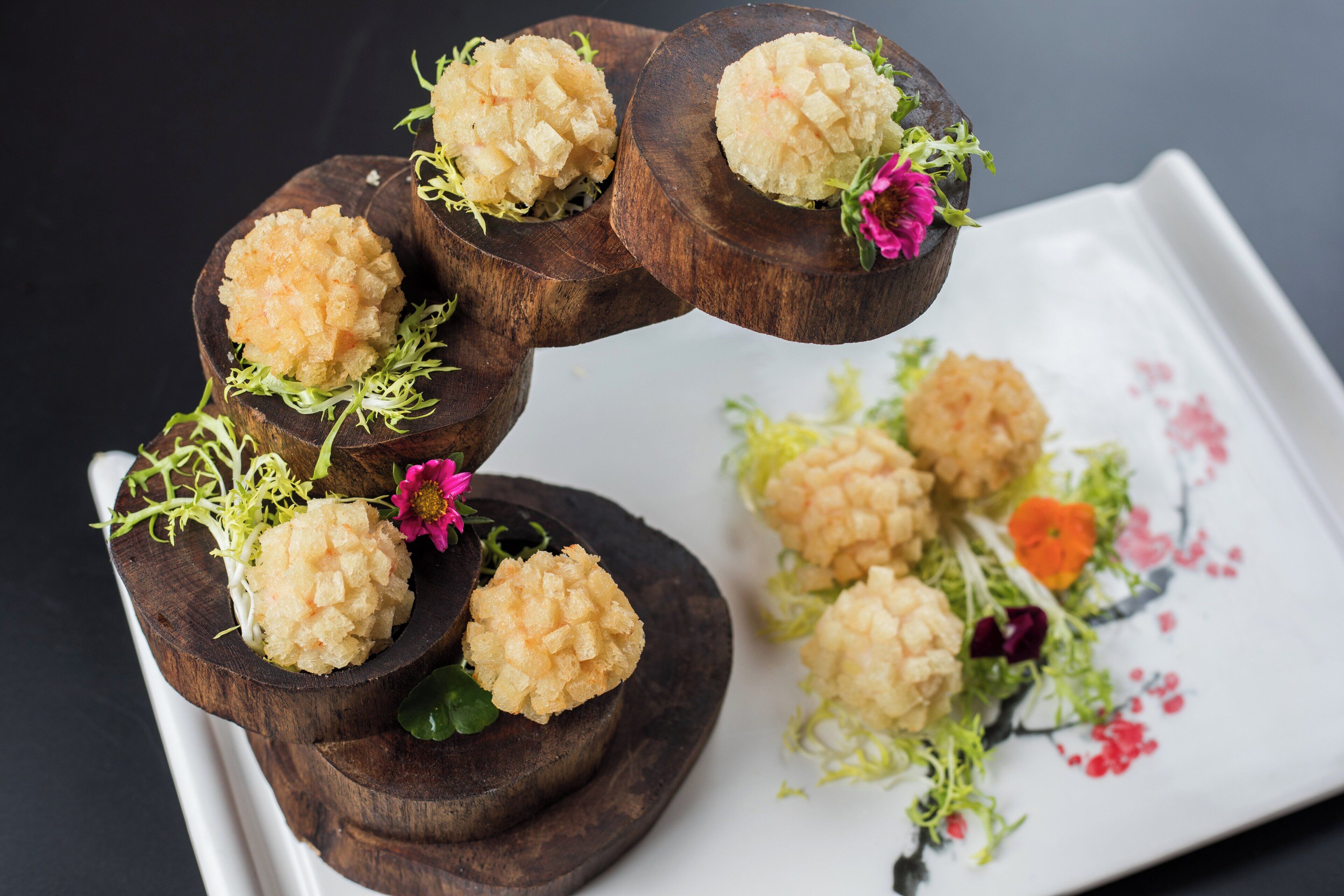 Una tabla de madera con alimentos esféricos y un arreglo floral sobre un plato blanco.