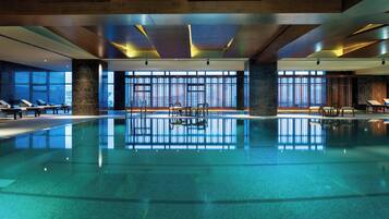 A large indoor swimming pool with clear blue water, surrounded by lounge chairs and a modern architectural design with large windows.