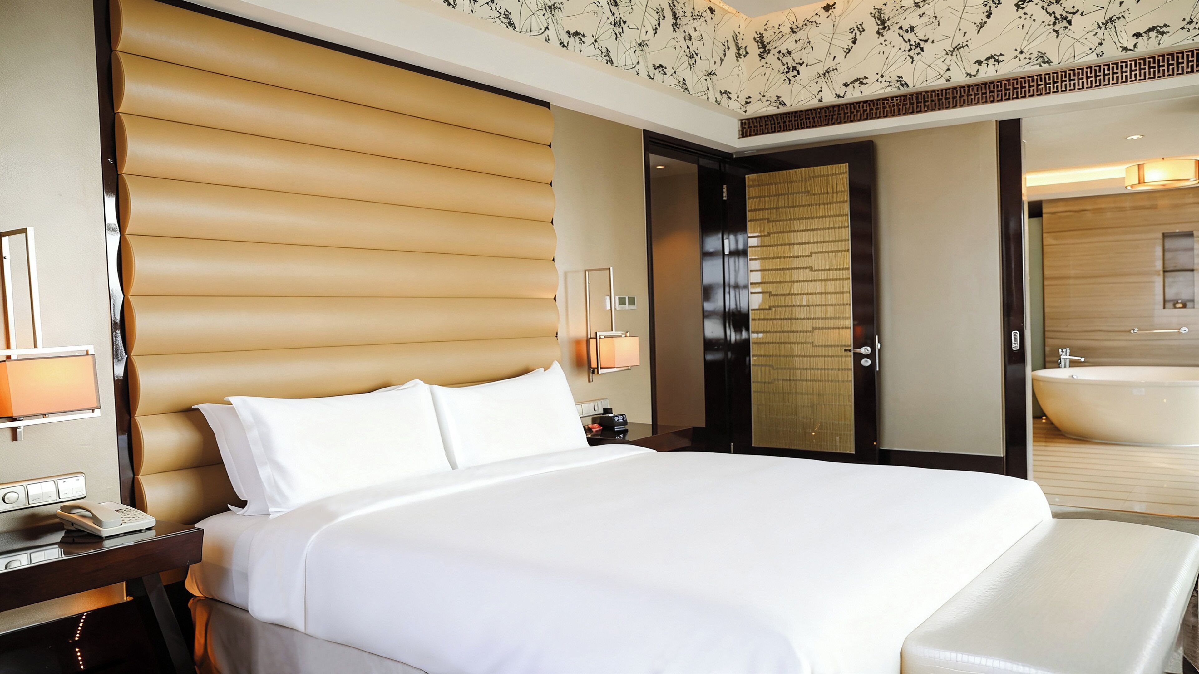 A hotel room with a large bed, bedside table, lamp, and a bathroom with a bathtub.