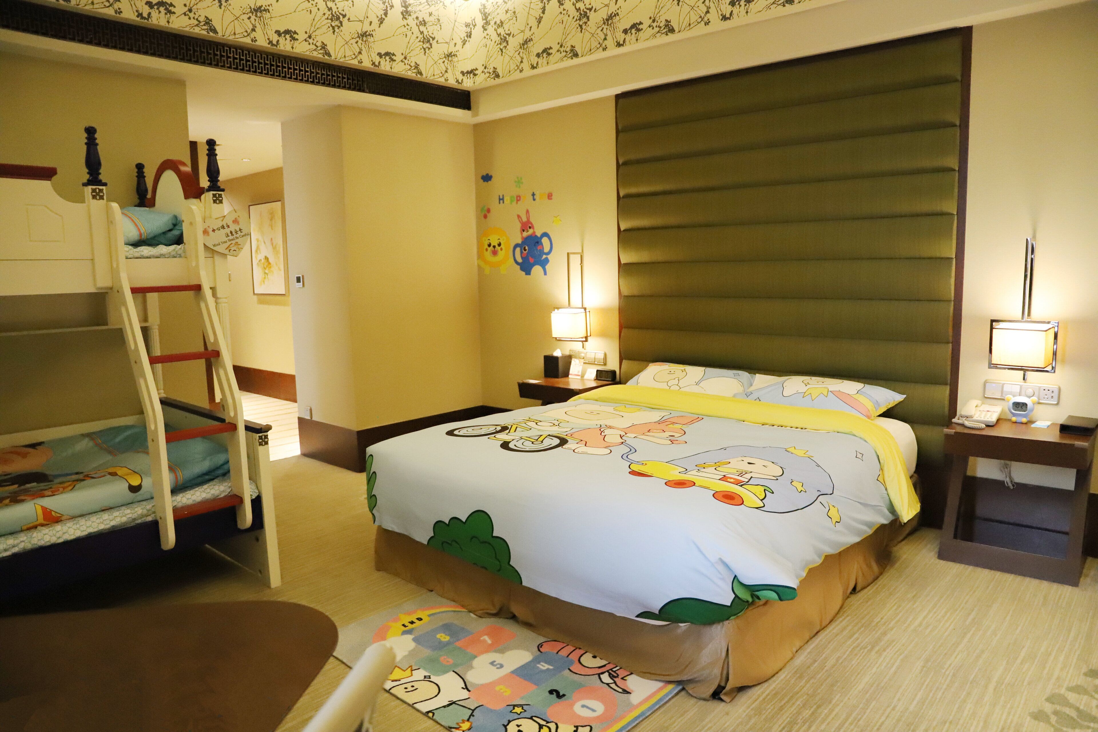 Una habitación de hotel con literas, una cama grande con colcha de diseño infantil, dos mesitas de noche con lámparas y una pequeña mesita auxiliar.