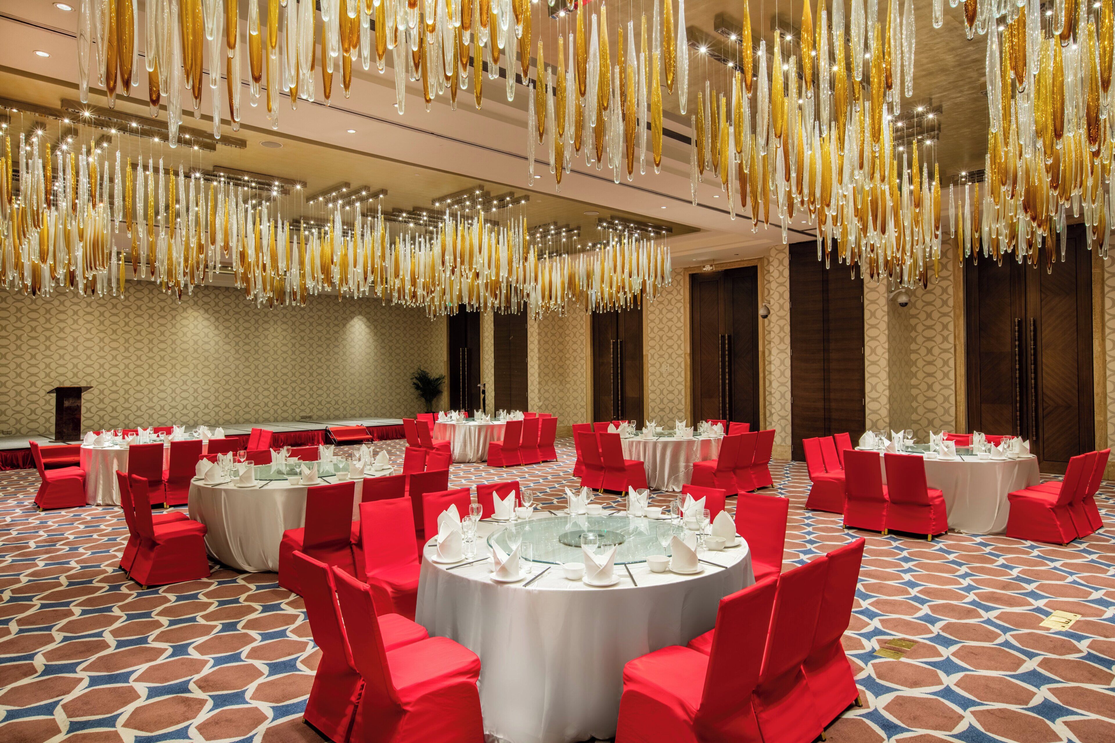 A banquet hall with round tables, red chairs, and hanging crystal chandeliers.