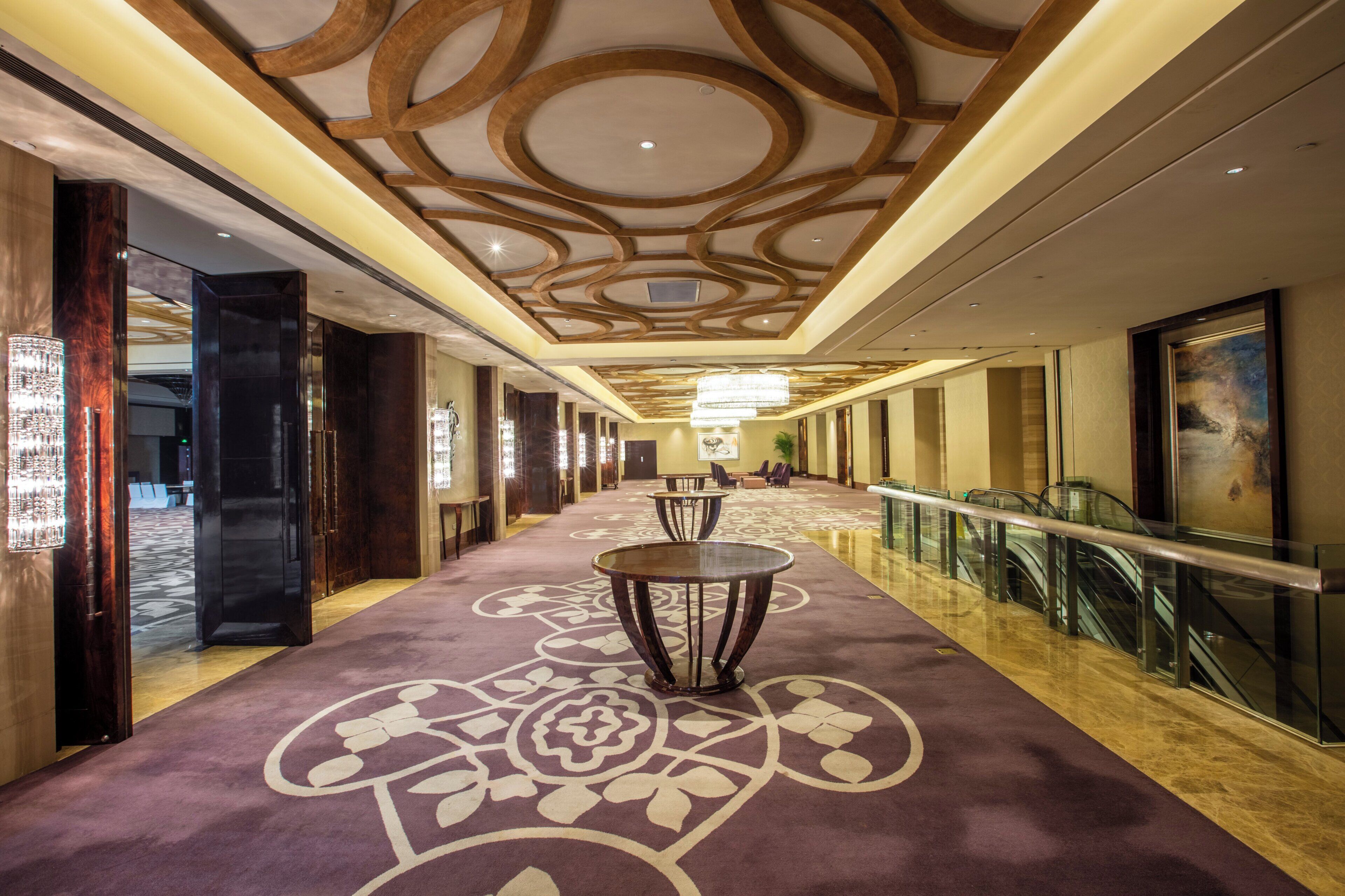 A hotel corridor with a patterned carpet, a chandelier, and a glass railing.