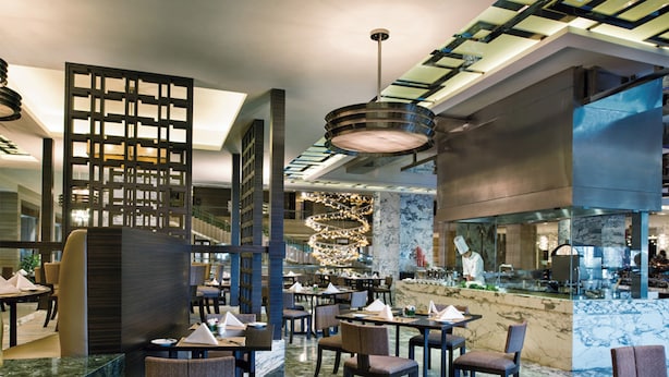 A modern restaurant with marble flooring, dark-toned chairs, and a central chandelier.