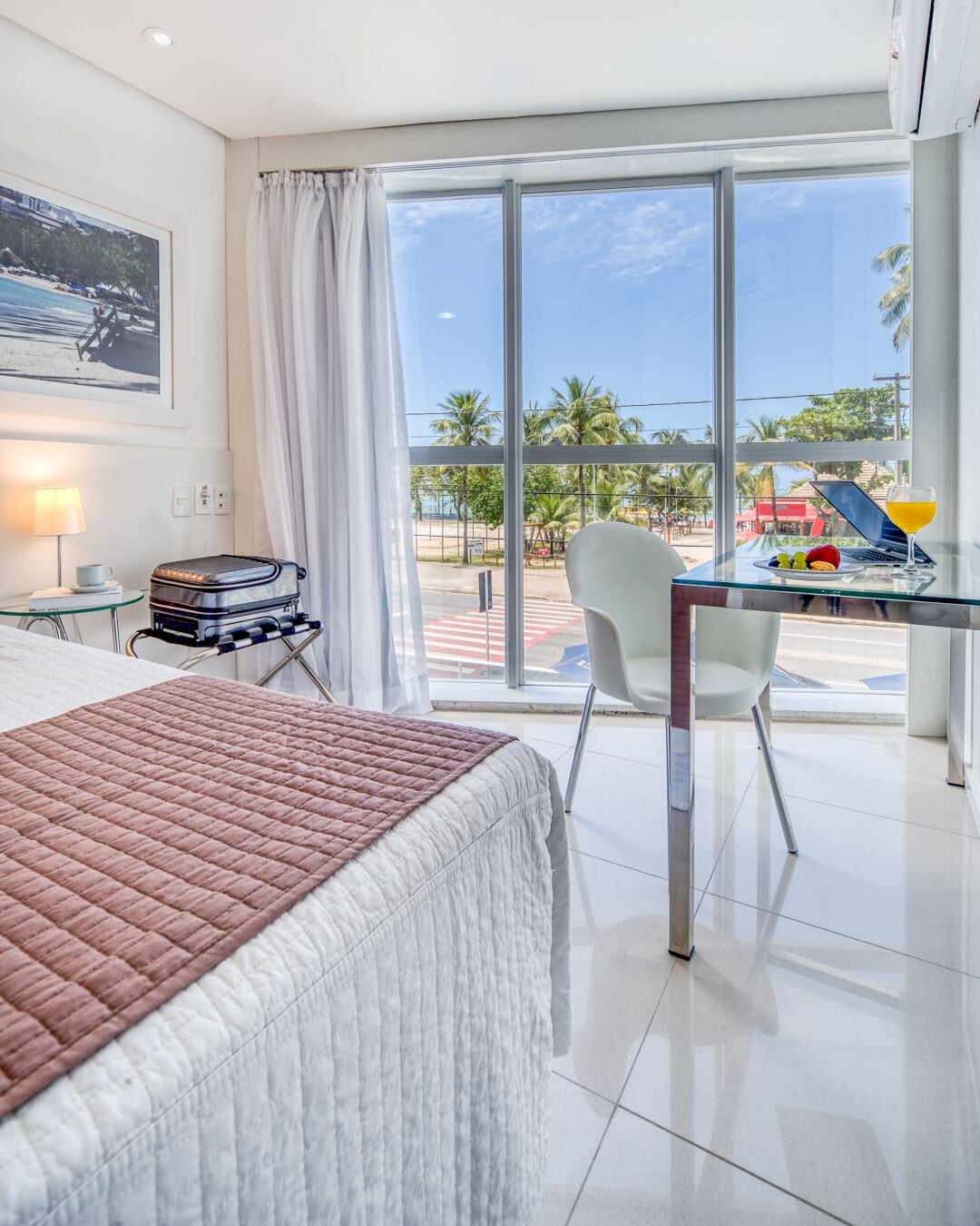 A hotel room with a bed, a desk with a laptop and a drink, a suitcase, and a view of a beach and palm trees.