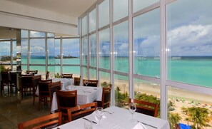 Un restaurante con amplios ventanales que ofrece vistas a una playa y al océano. Las mesas están dispuestas con manteles blancos y copas de vino.