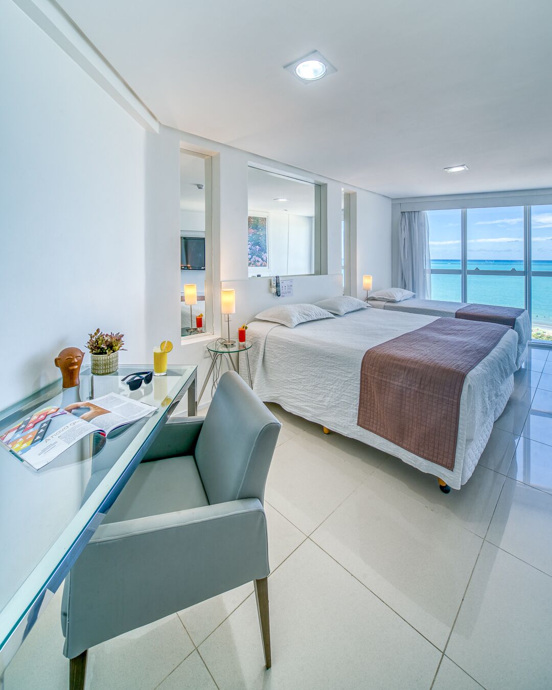 Quarto de hotel moderno com uma cama grande, uma escrivaninha com cadeira, uma mesa de vidro e vista para o oceano.