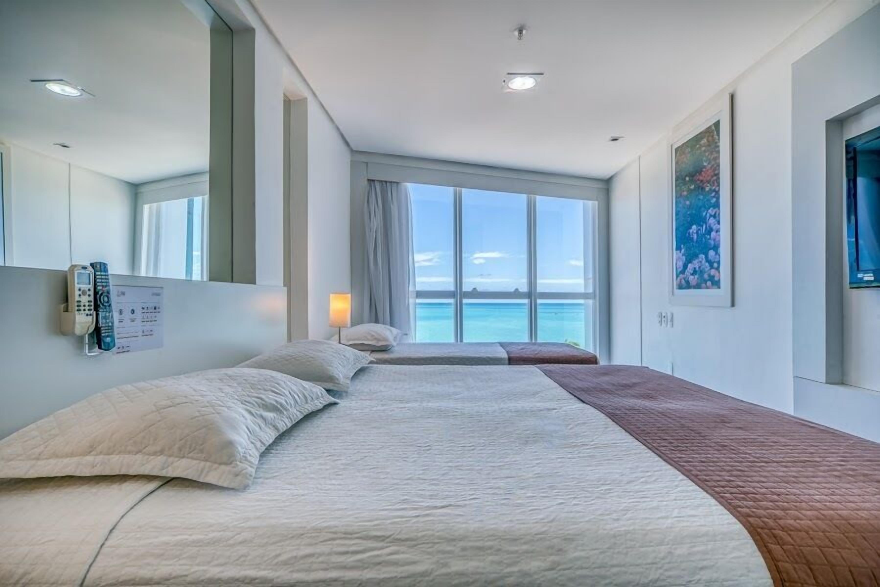 A hotel room with a large bed, a bedside table, a lamp, a phone, and a window with a view of the sea.