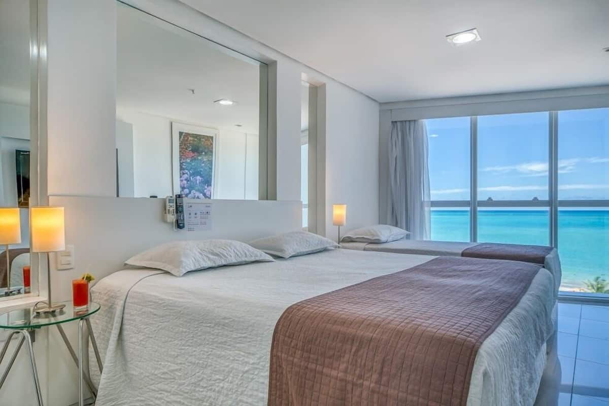 A hotel room with a large bed, a bedside table, a mirror, and a view of the ocean.