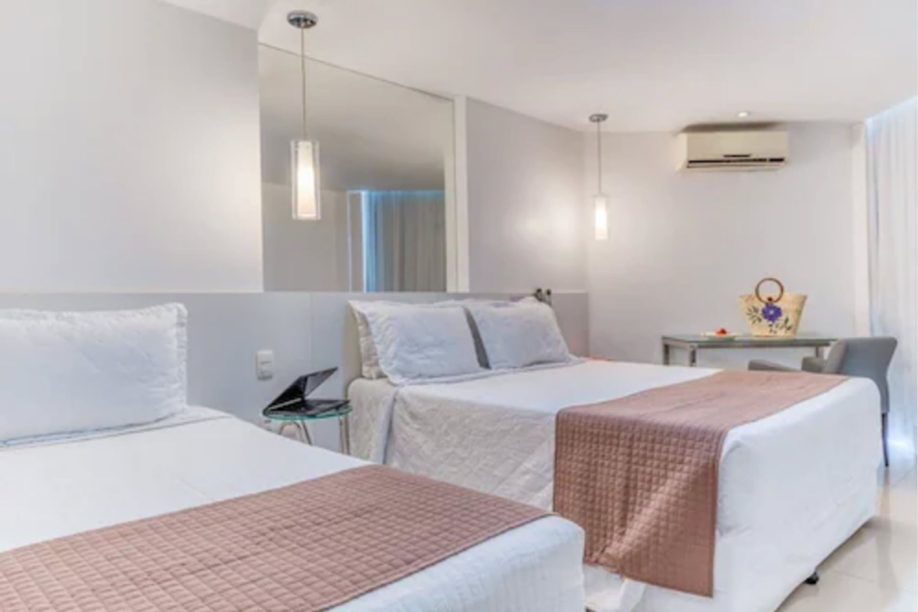 A hotel room with two beds, a small table, a chair, a mirror, and an air conditioning unit.