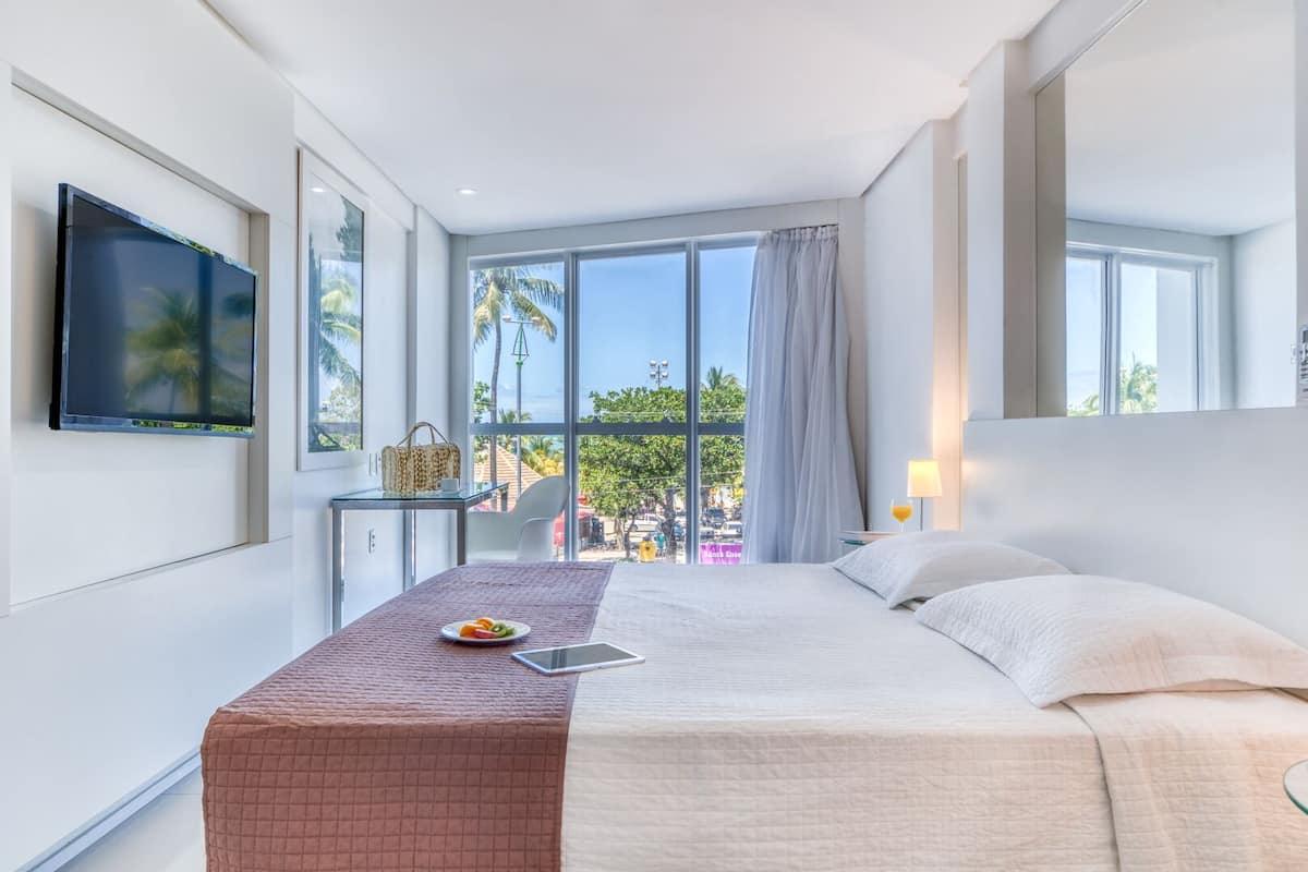 A hotel room with a bed, a desk, a TV, and a view of palm trees and a beach.