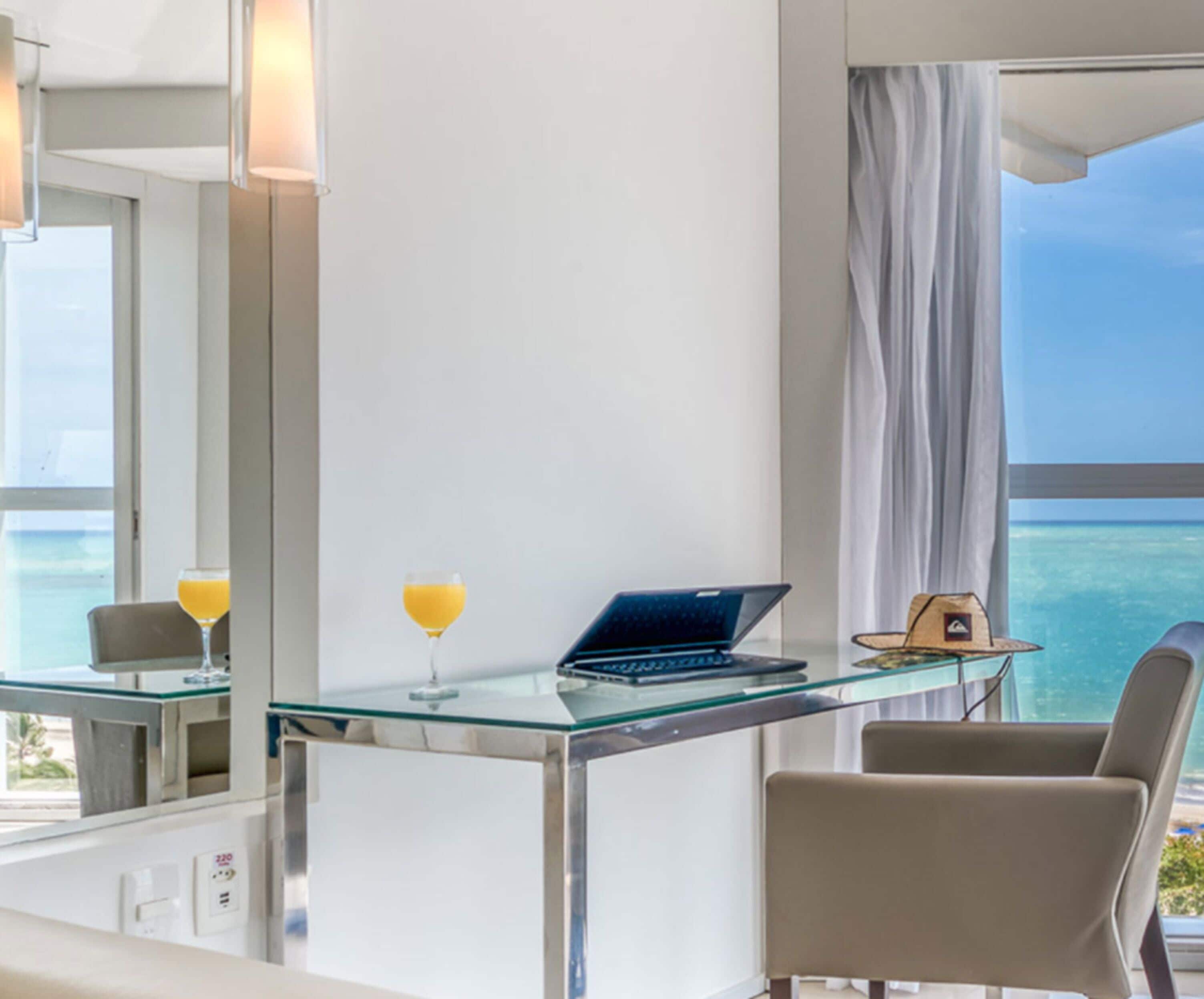 A modern room with a glass desk, laptop, two drinks, and a view of the ocean.