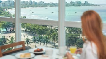 Vista desde una ventana sobre una playa con palmeras, un horizonte urbano y barcos en el agua.