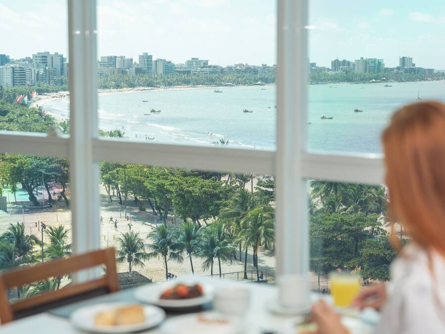 Vista desde una ventana sobre una playa con palmeras, un horizonte urbano y barcos en el agua.