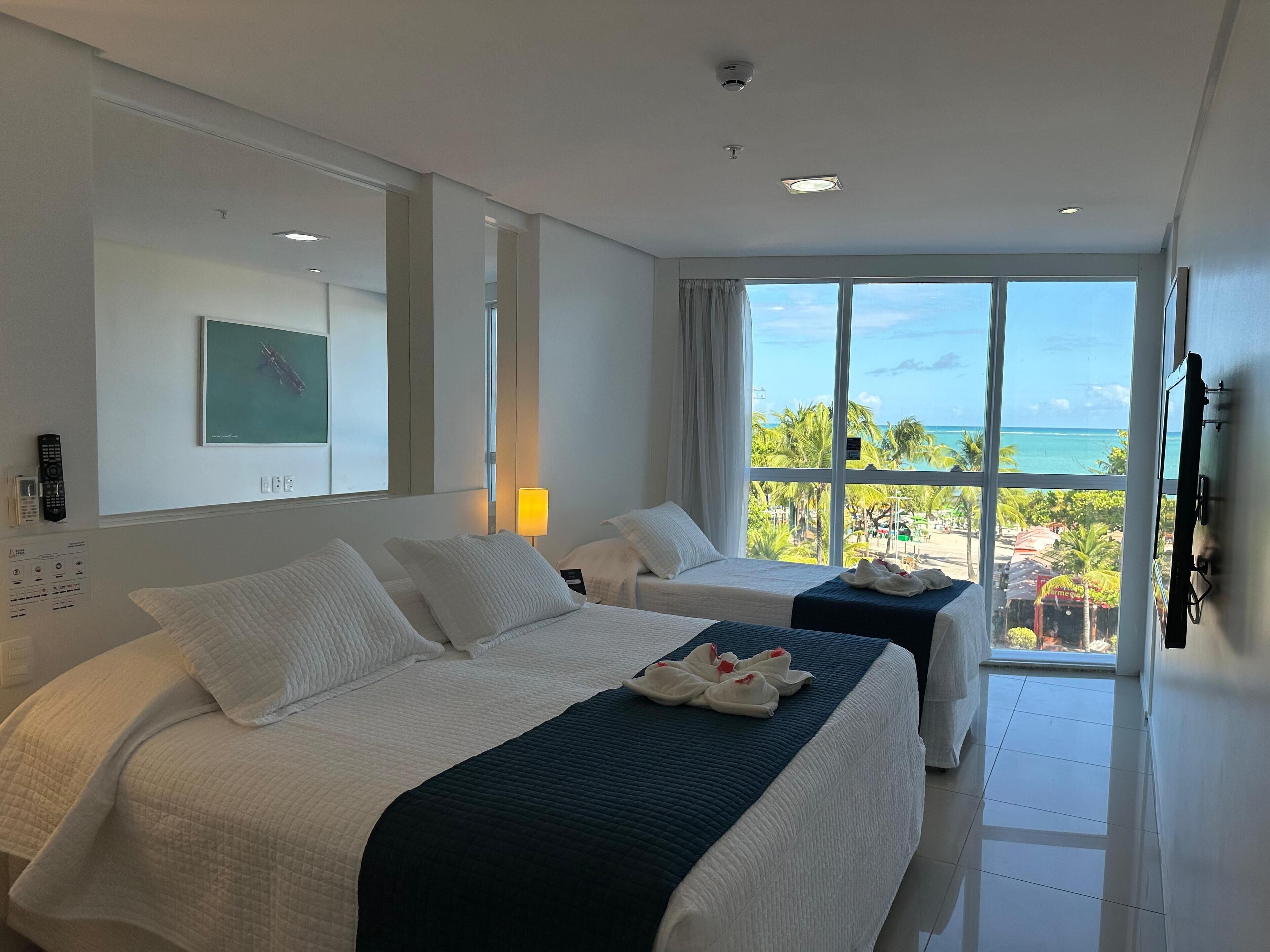 Habitación de hotel con dos camas, un ventanal con vista a la playa y un televisor montado.