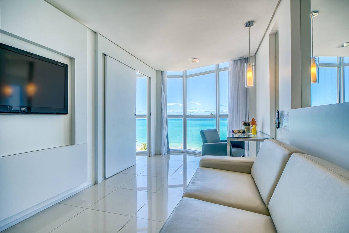A modern hotel room with a large window offering a view of the sea, a flat-screen TV mounted on the wall, a sofa, and a small table with a drink.