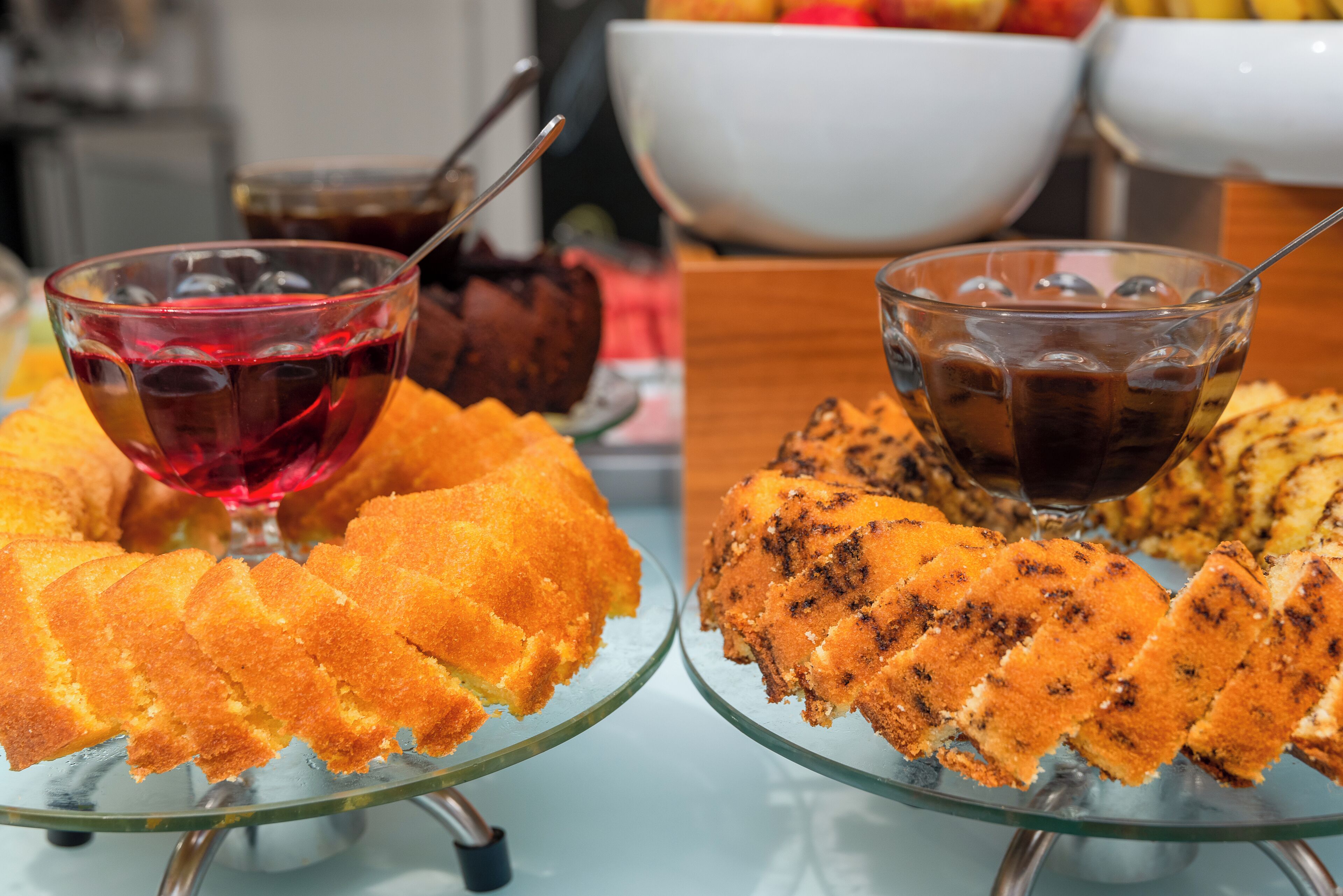 Two glass cake stands with sliced cakes and two glasses of iced beverages on a table.