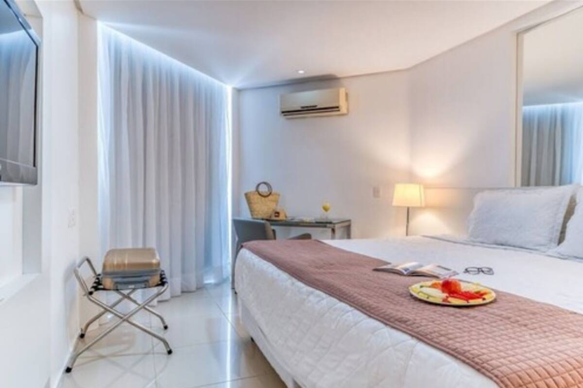 A hotel room with a bed, a suitcase, a chair, a table with a lamp, and an air conditioning unit.