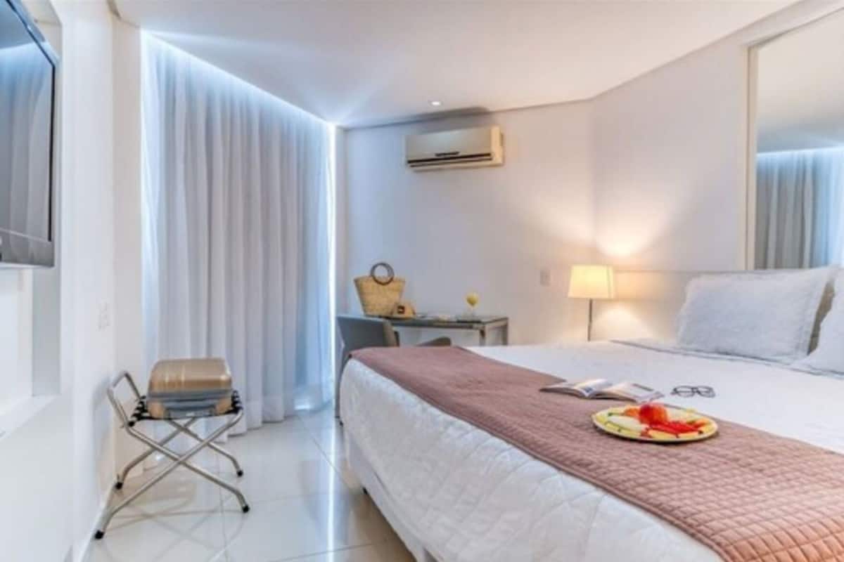 A hotel room with a bed, a desk, a chair, a suitcase, and an air conditioning unit.