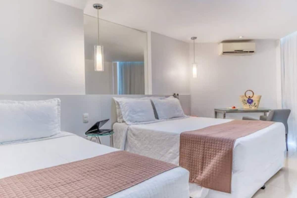 A hotel room with two beds, a small table, a chair, a mirror, and an air conditioning unit.