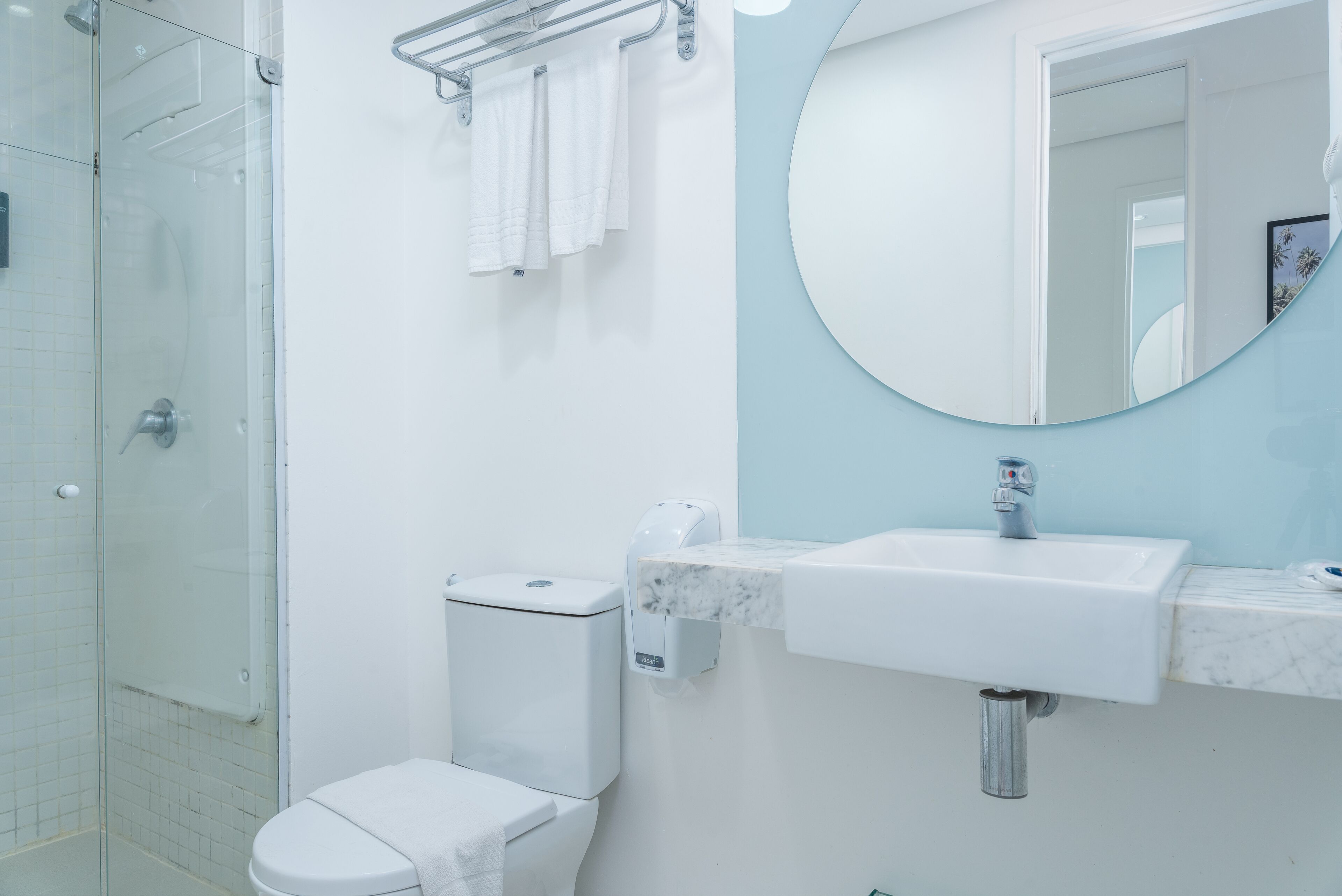A modern bathroom with a glass shower enclosure, a white toilet, a sink with a marble countertop, and a round mirror.