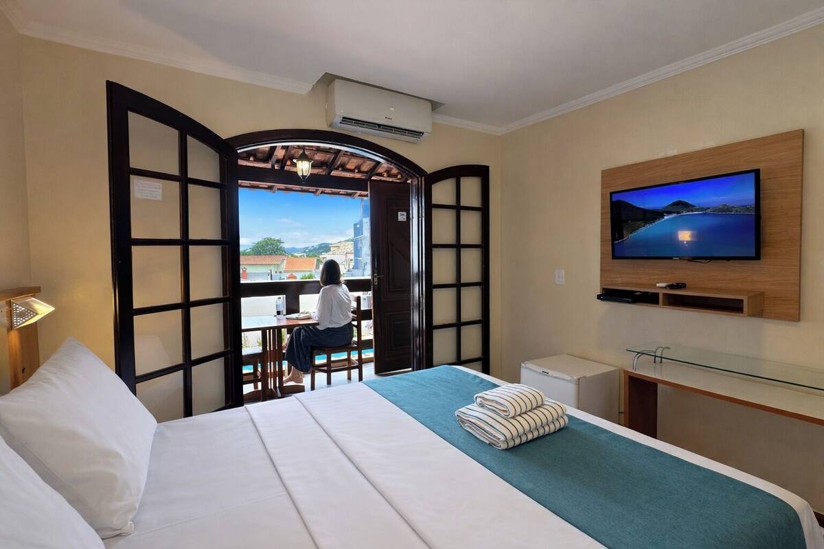 A hotel room with a bed, a TV mounted on the wall, a window with a view of buildings, and a door leading outside.